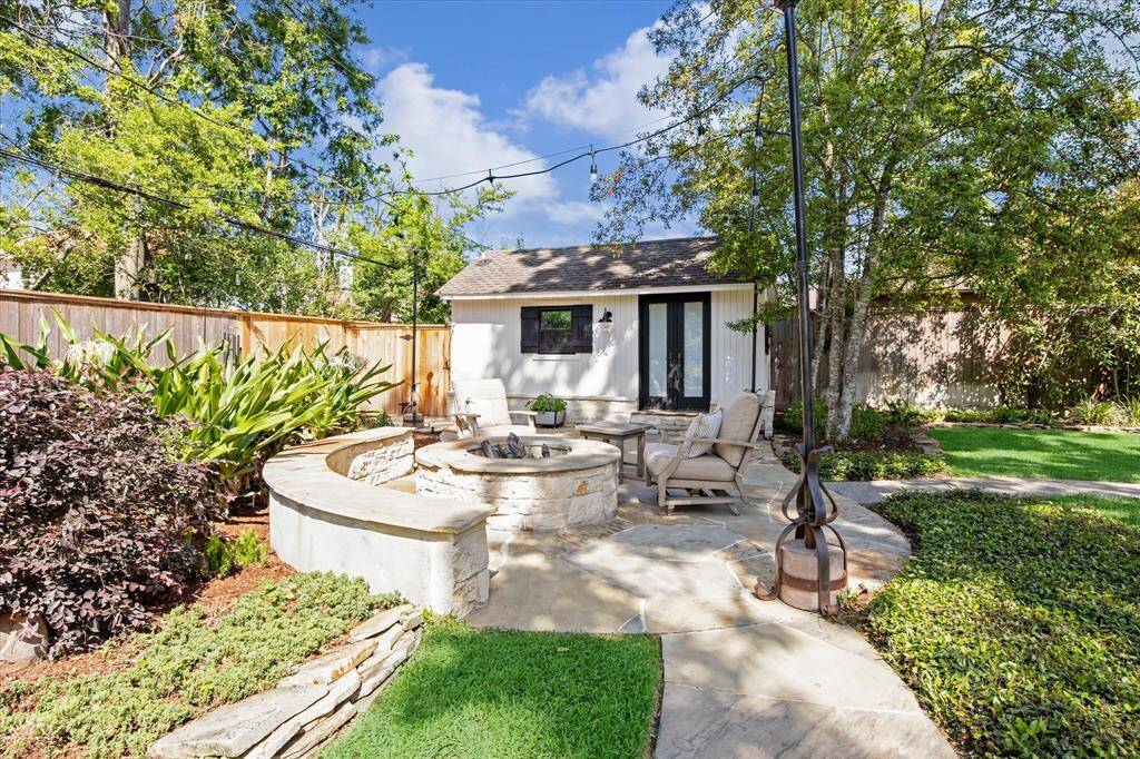 This photo features a charming casita with full bathroom in a backyard setting. It has white siding, black shutters, and French glass doors. The area is surrounded by mature trees and enclosed by a wooden fence, with a stone patio and fire pit providing a quaint outdoor space.
