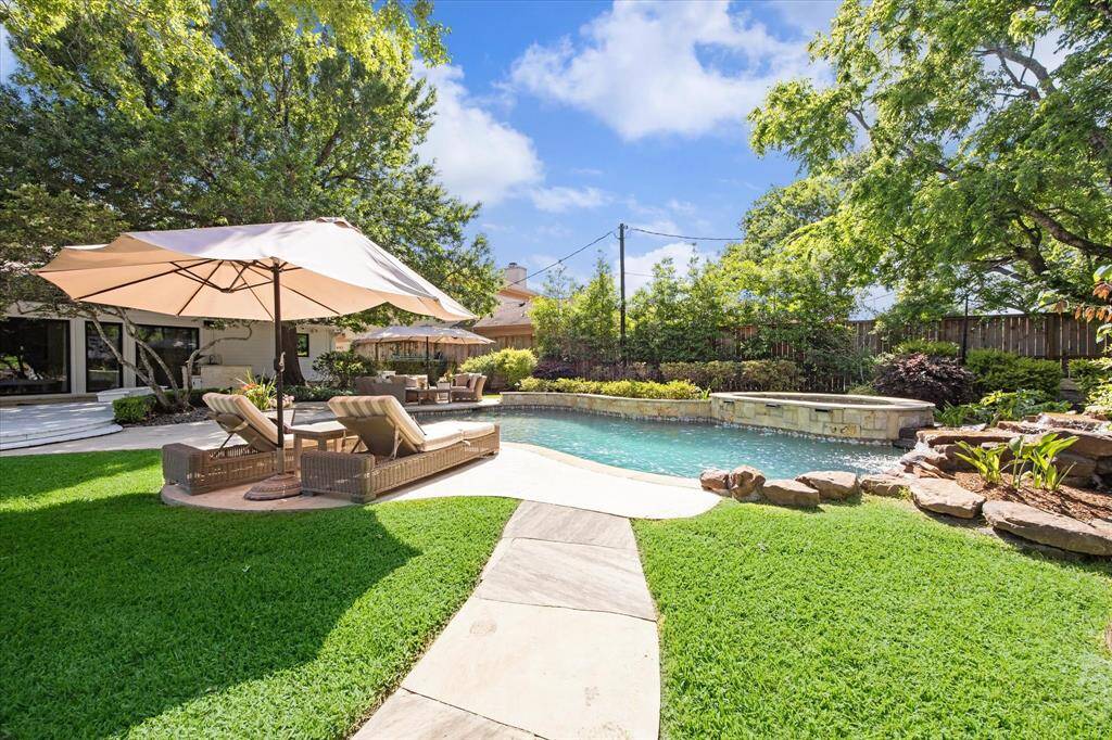 This backyard features a beautiful swimming pool with a stone border, surrounded by lush greenery and mature trees. There's a comfortable seating area with lounge chairs and umbrellas on a patio, offering a perfect spot for relaxation or entertaining.