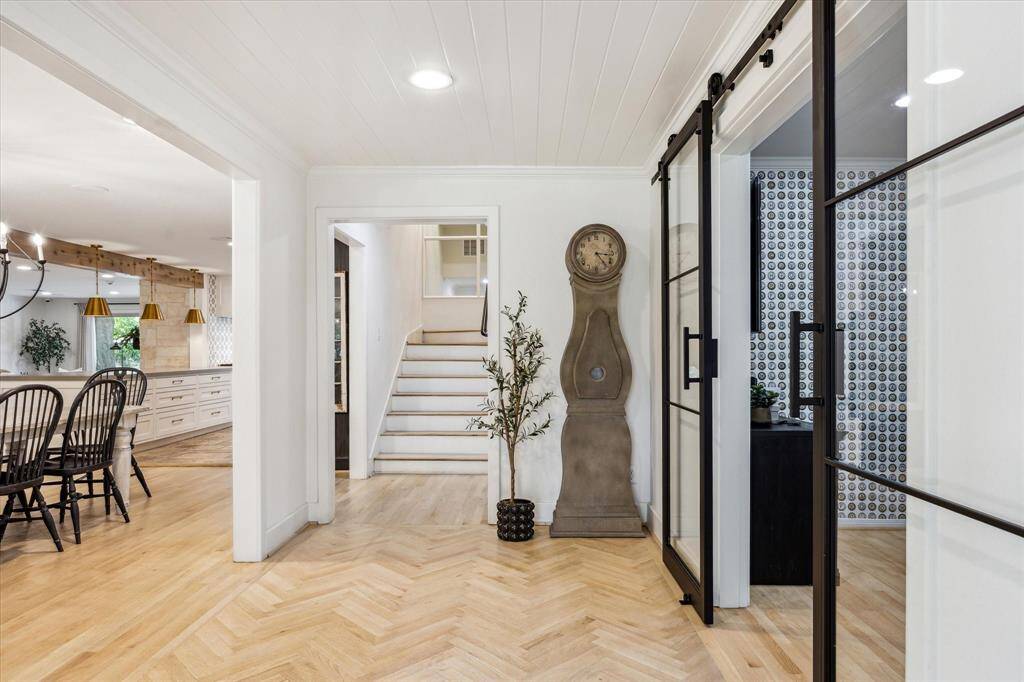 This photo showcases a bright, inviting entryway with light wood herringbone flooring and a staircase leading to an upper level.  The adjacent dining area has elegant lighting and seating, creating a warm and welcoming atmosphere.