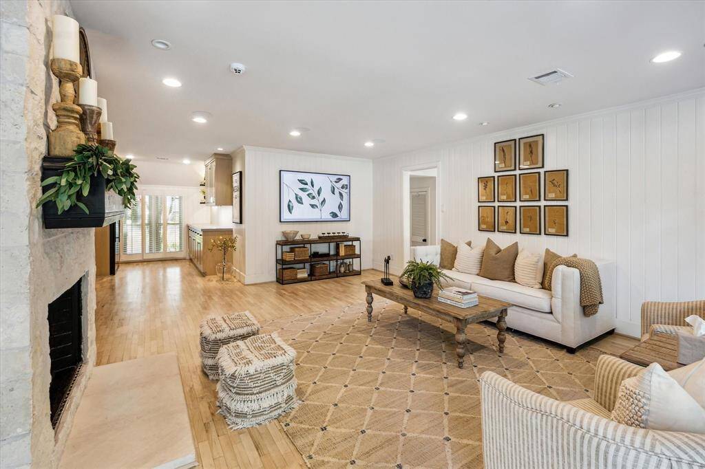 This inviting living room features hardwood floors, a cozy stone fireplace, and a bright, open layout. The room flows seamlessly into a well-lit kitchen area, perfect for entertaining.