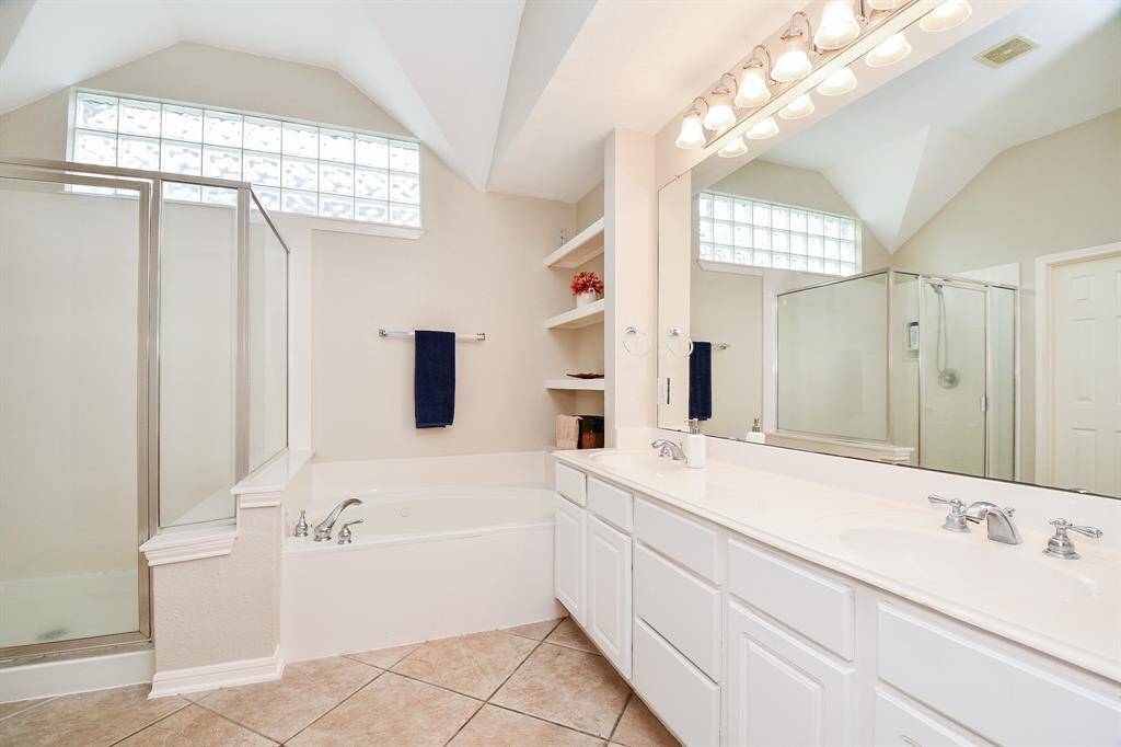 There's just something about a primarily light colored bathroom that soothes us, like this primary ensuite bath; with dual sinks nestled in a long, sleek counter, beneath a shining wall-wide mirror and delicate lighting, above. Note the built-in shelving treatment to the right of soaking tub and walk-in shower.