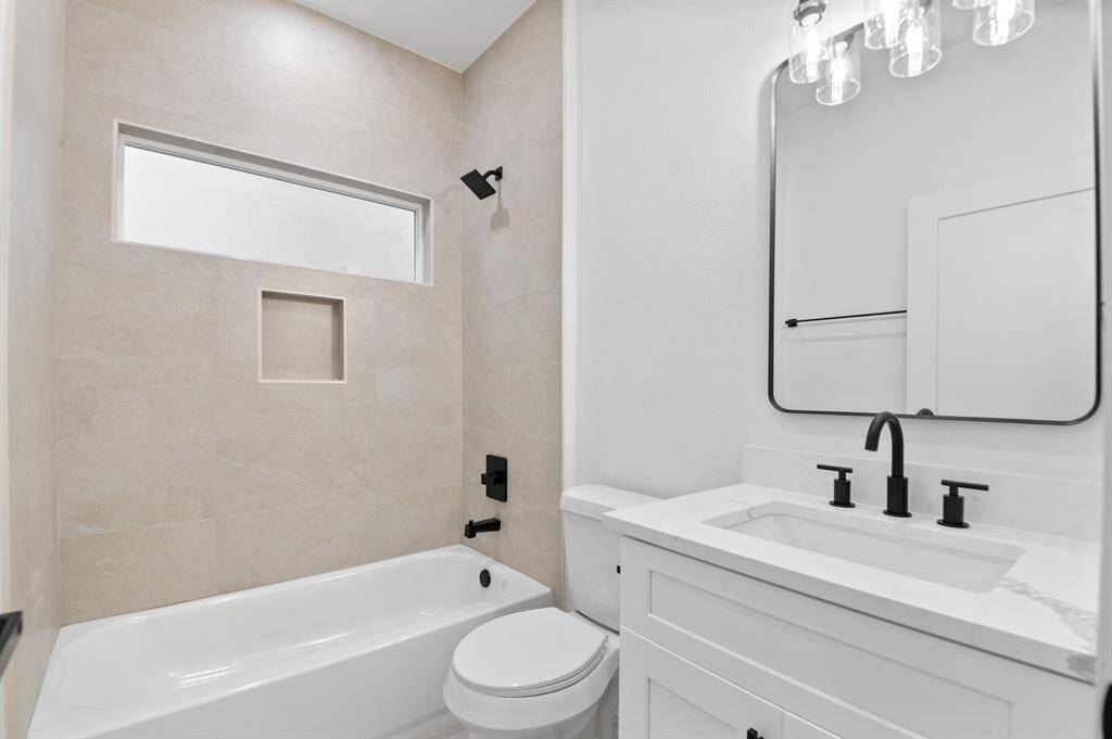 This modern bathroom features a sleek design with a tiled bathtub, a built-in shelf, and a frosted window for natural light. The vanity offers a rectangular mirror, stylish black fixtures, and a clean white countertop.