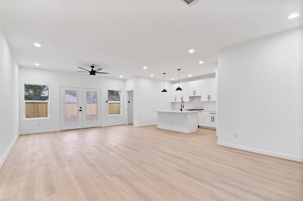 This photo showcases a spacious, open-concept living area with light wood flooring, a modern kitchen featuring a central island and pendant lighting, and large windows for natural light. The room is well-lit with recessed lighting and has a ceiling fan for comfort.