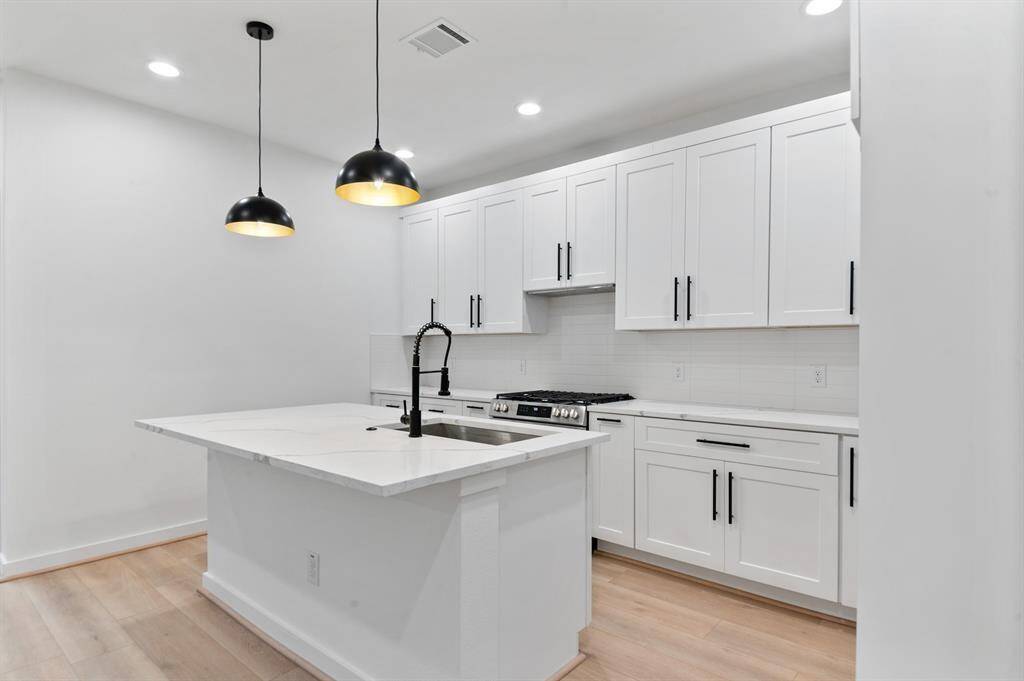 Modern kitchen with white cabinetry, a large island with a sink, and stylish black pendant lighting. Features ample storage, stainless steel appliances, and light wood flooring for a clean, contemporary look.