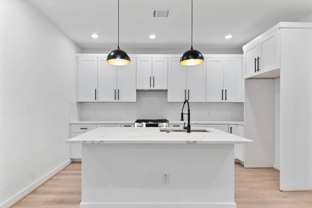 This modern kitchen features a sleek design with white cabinetry, a spacious island with a built-in sink, and stylish black pendant lights. The light wood flooring and minimalist aesthetic create a bright and open feel.