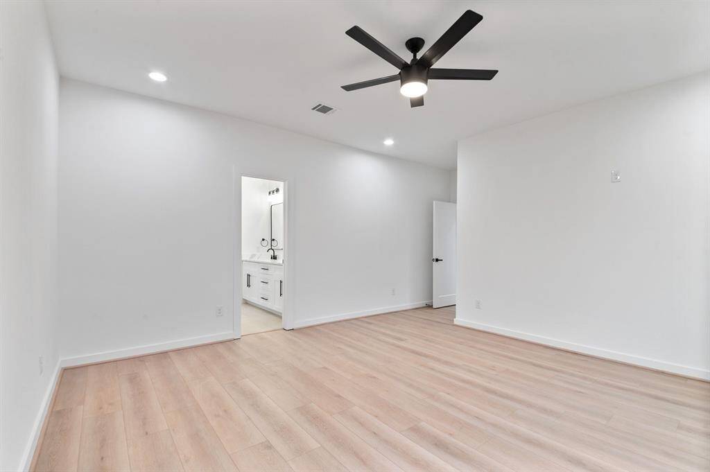 This photo showcases a spacious, modern bedroom with light wood flooring and recessed lighting. It features a ceiling fan and leads to an en-suite bathroom with a sleek vanity and dual sinks.