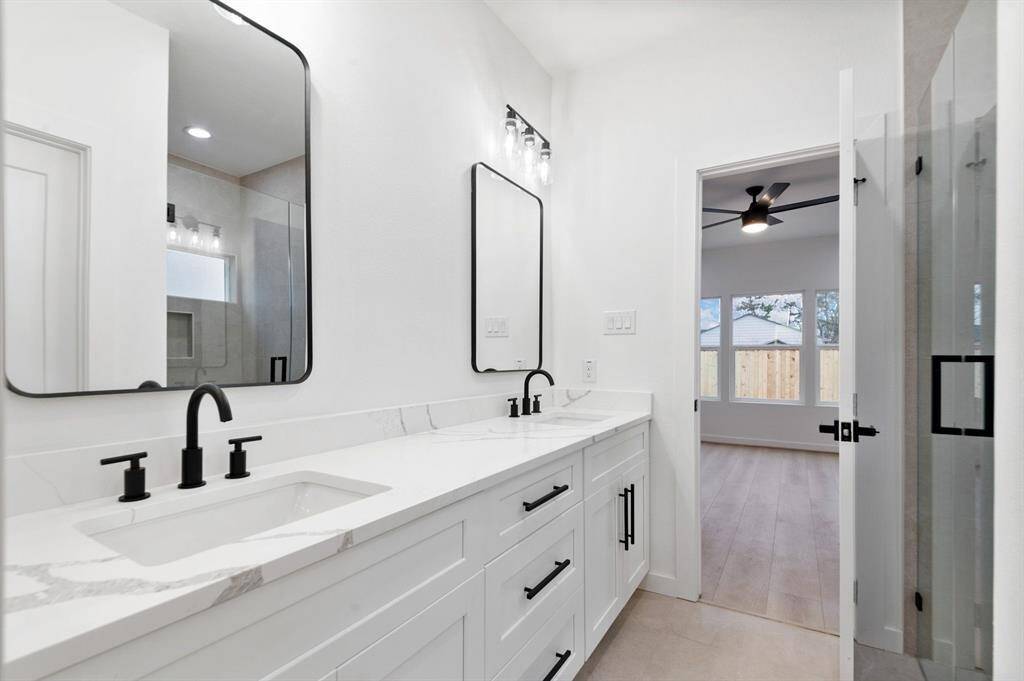 This photo showcases a modern bathroom with a double vanity featuring sleek black fixtures and large mirrors. The room is bright with white walls and a view into an adjacent room with a ceiling fan and large windows.