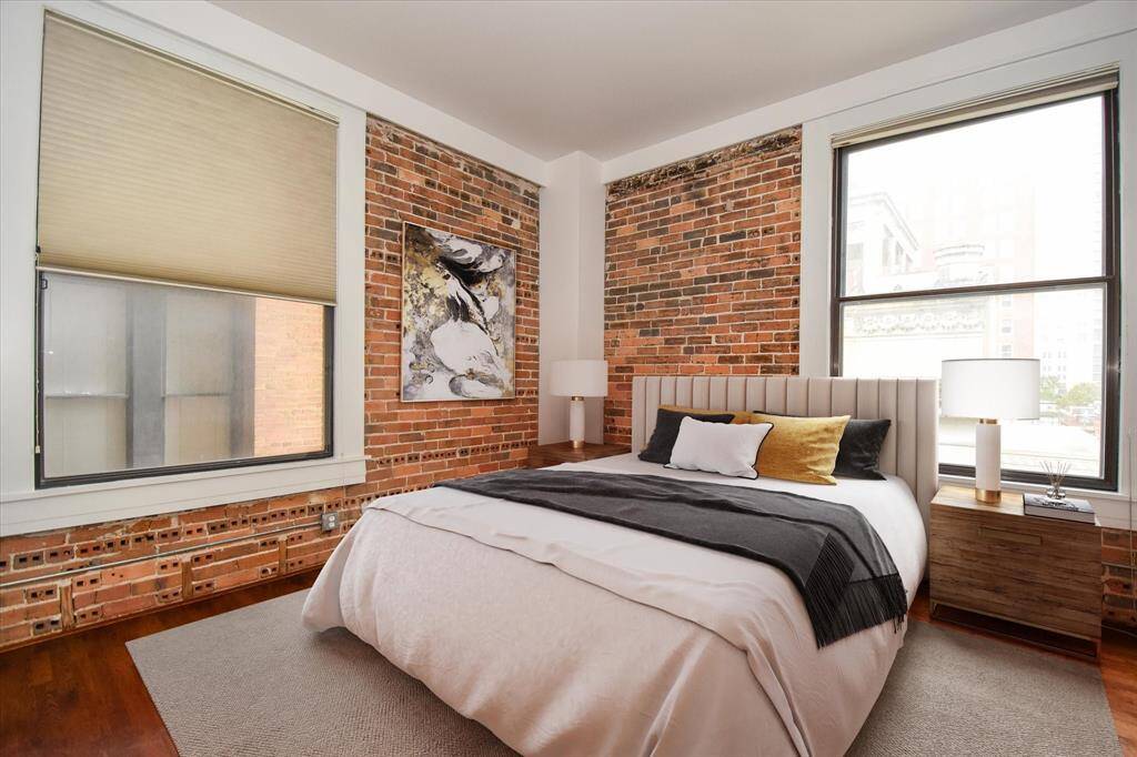 This inviting bedroom features classic exposed brick, large windows for natural light, and a modern design with warm wood floors, perfect for downtown living.