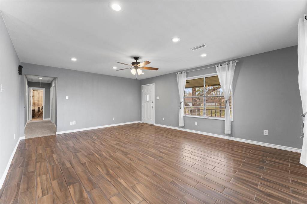 This spacious living room features wood-look tile flooring, neutral gray walls, and ample natural light from large windows with white curtains. It includes a ceiling fan and recessed lighting, creating a bright and airy atmosphere.