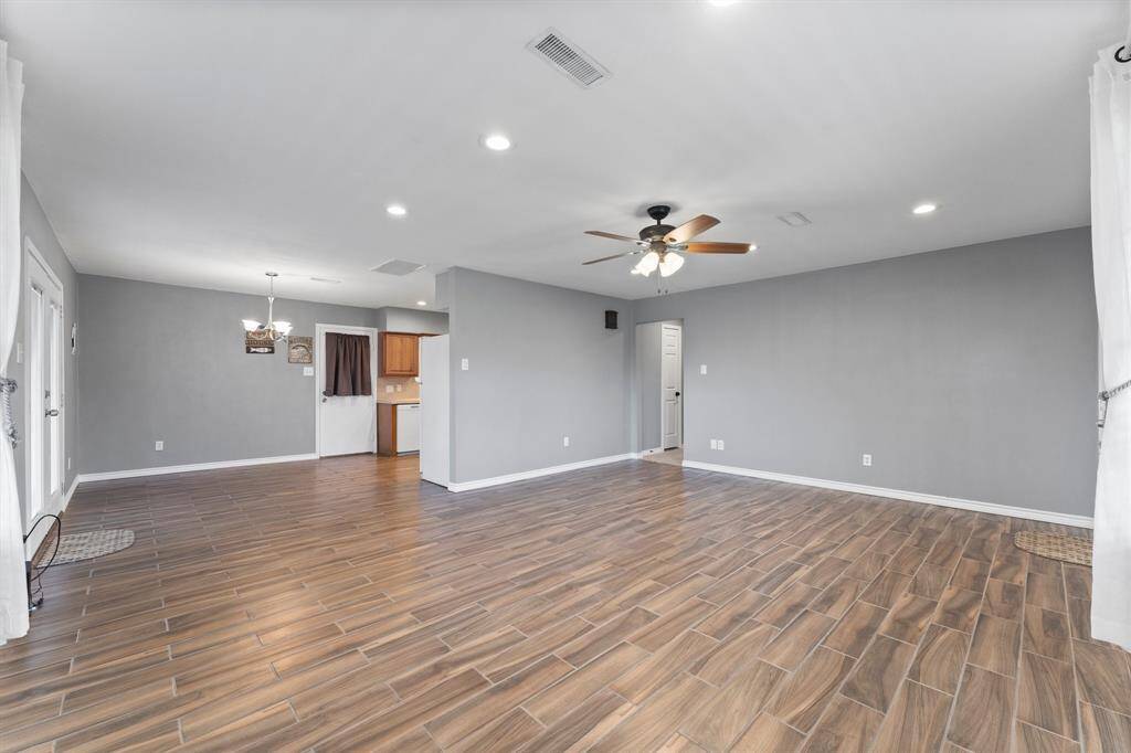 Bright and spacious living room with wood-look tile flooring, neutral gray walls, recessed lighting, and a ceiling fan. Large window with sheer curtains offers natural light and a view of the outdoors.