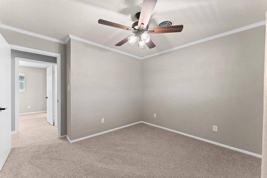 This bedroom features neutral walls and carpet, a ceiling fan with lights, and a window with curtains, providing a cozy space with plenty of natural light.