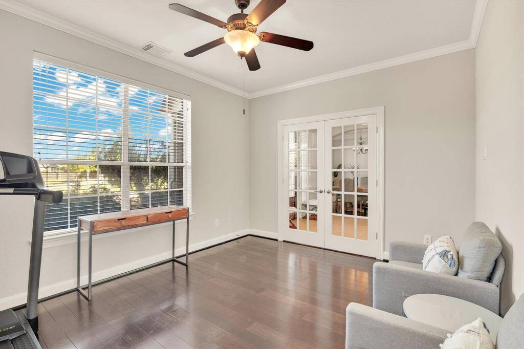 This bright room features dark hardwood floors and a ceiling fan, with large windows offering plenty of natural light. French doors open to an adjoining space, creating an inviting and versatile area for an office or sitting room.