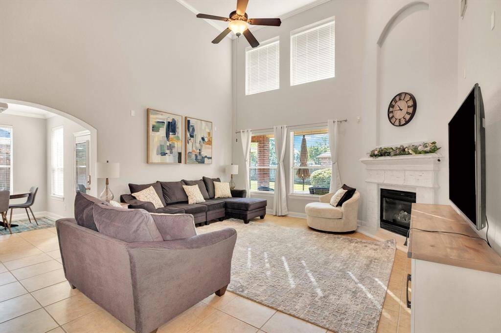 This spacious living room features a high ceiling with large windows for ample natural light. It includes a cozy fireplace, modern decor, and a comfortable sectional sofa. The open layout flows into a dining area, enhancing the home's inviting atmosphere.