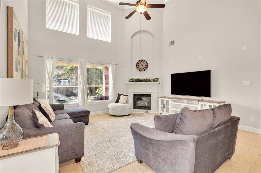 Bright, spacious living room with high ceilings and large windows. Features a cozy fireplace, modern ceiling fan, and neutral tones for a welcoming atmosphere. Ideal for relaxing and entertaining.