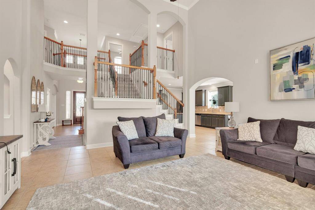 This spacious living room features high ceilings, a modern open staircase with wooden accents, and a bright, neutral color palette. It flows seamlessly into a kitchen area, creating an inviting open-concept space perfect for entertaining.