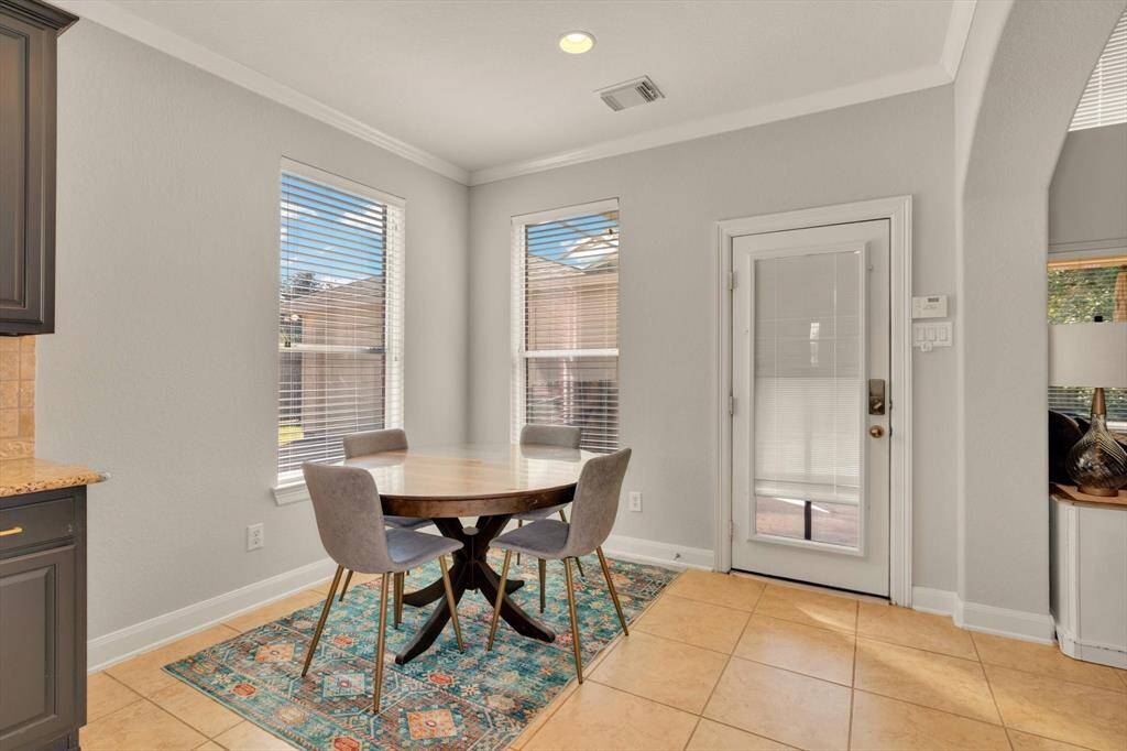 Bright breakfast area with tile flooring, featuring a round table and four chairs. Large windows and a glass door provide plenty of natural light, opening to a pleasant outdoor view. Neutral walls and a colorful area rug add charm.
