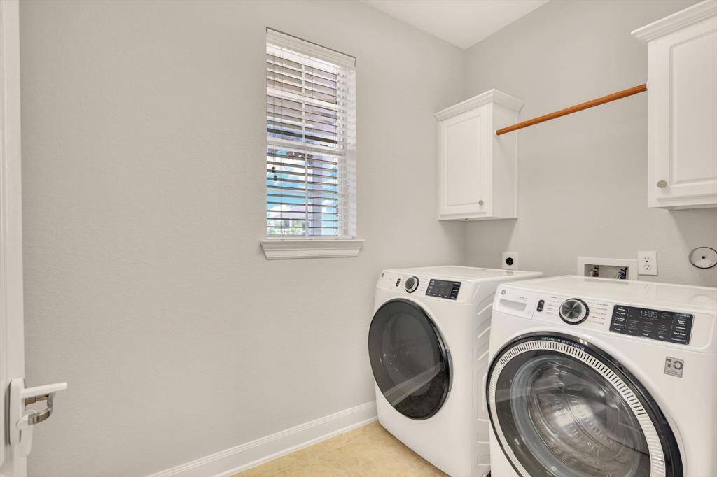 This is a bright, modern laundry room featuring a washer and dryer set, white cabinetry for storage, a hanging rod, and a window for natural light.