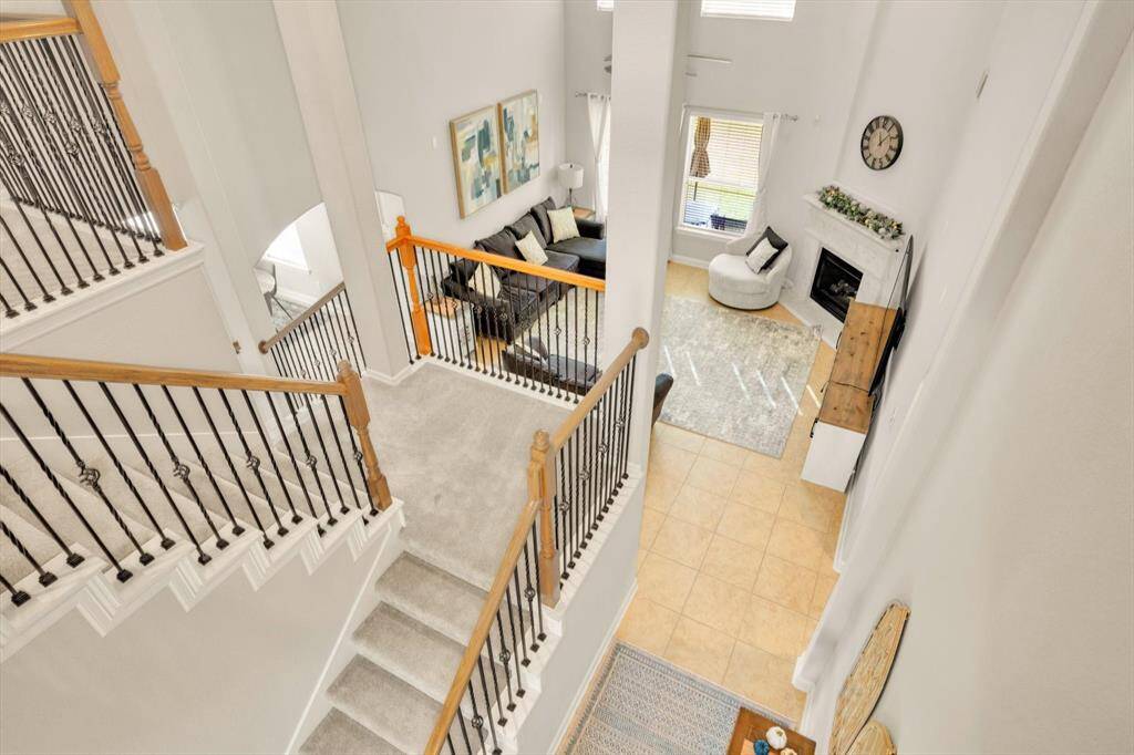 A spacious, open-concept living area with high ceilings. The view from the staircase highlights a cozy living room featuring a fireplace, large windows, and a blend of carpet and tile flooring. Stylish railings and modern decor enhance the inviting atmosphere.