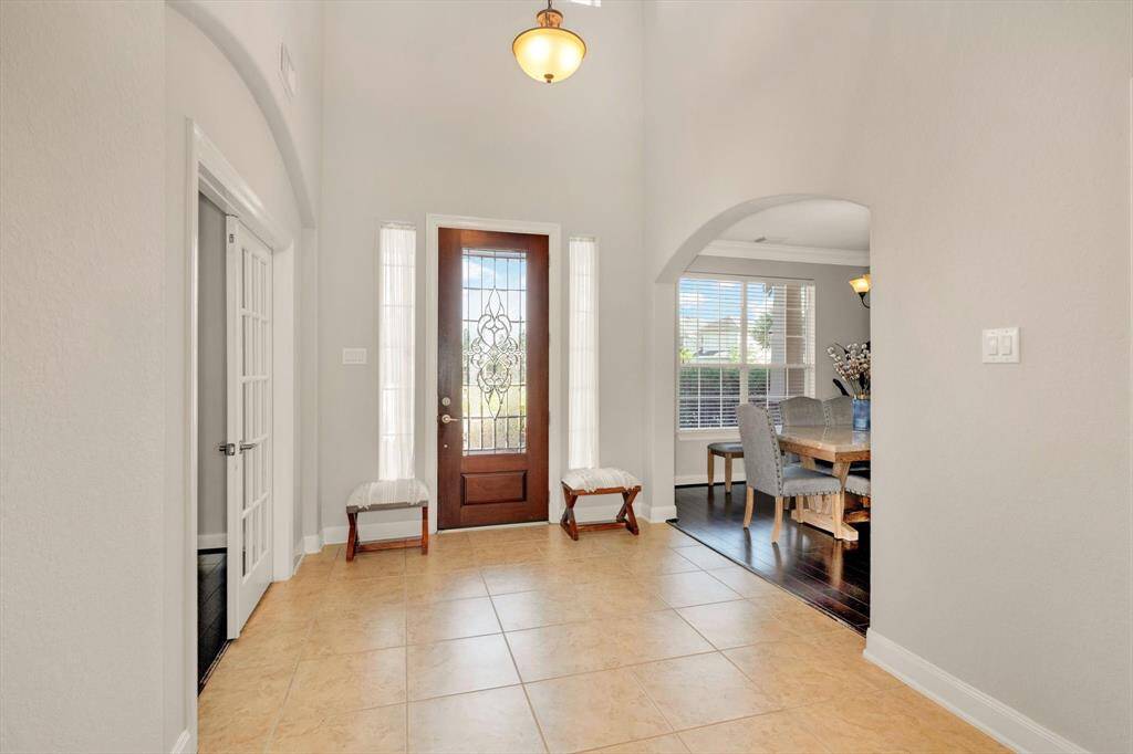 Bright entryway with high ceilings and tile flooring, featuring a wooden front door with glass panels. To the right, an open archway leads to a dining room with large windows and hardwood floors. A side door with frosted glass adds elegance.