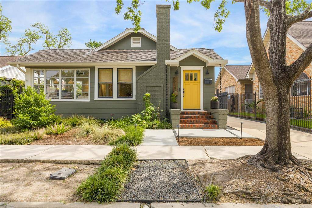 Charming single-story home with a cozy front porch, featuring a bold yellow door and brick steps. The exterior is painted in a muted green, complemented by well-maintained landscaping and mature trees. The large windows provide plenty of natural light, creating a welcoming atmosphere.