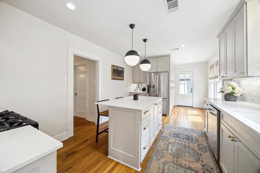 This kitchen features a bright, modern design with light cabinetry, stainless steel appliances, and a central island with seating. Pendant lights and open shelving add style, while a large window provides natural light. Perfect for both cooking and entertaining.