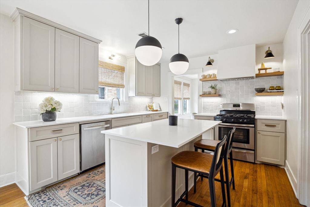 This kitchen features a bright, modern design with sleek cabinetry, stainless steel appliances, and a large central island with seating. It has warm hardwood floors, pendant lighting, and ample natural light, creating an inviting and functional space.