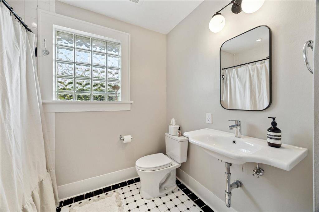 Bright bathroom with modern fixtures, featuring a large glass block window, a sleek white sink, and a wall-mounted mirror. The space includes a bathtub with a shower curtain and a stylish black-and-white tile floor.