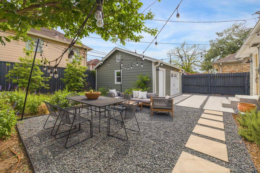 Charming backyard with a modern patio area, featuring string lights, a dining table, and cozy seating. The space is surrounded by greenery and has a sleek pathway leading to a small outbuilding, perfect for outdoor entertaining.