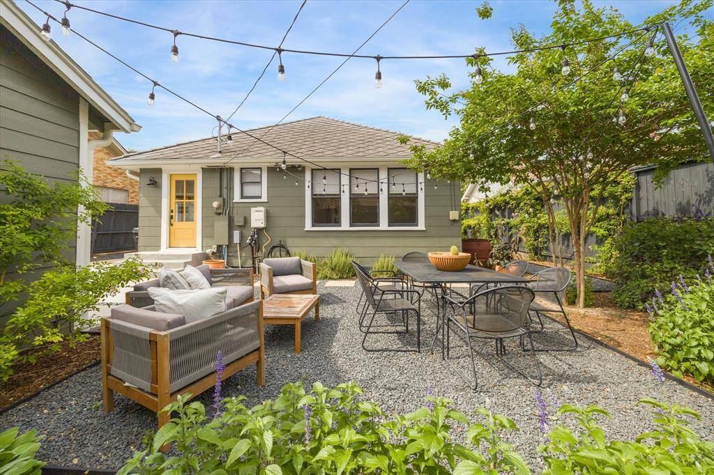 This inviting backyard features a cozy seating area with a sofa set and a dining table, surrounded by lush greenery. String lights add charm, creating a perfect outdoor space for relaxation and entertaining.