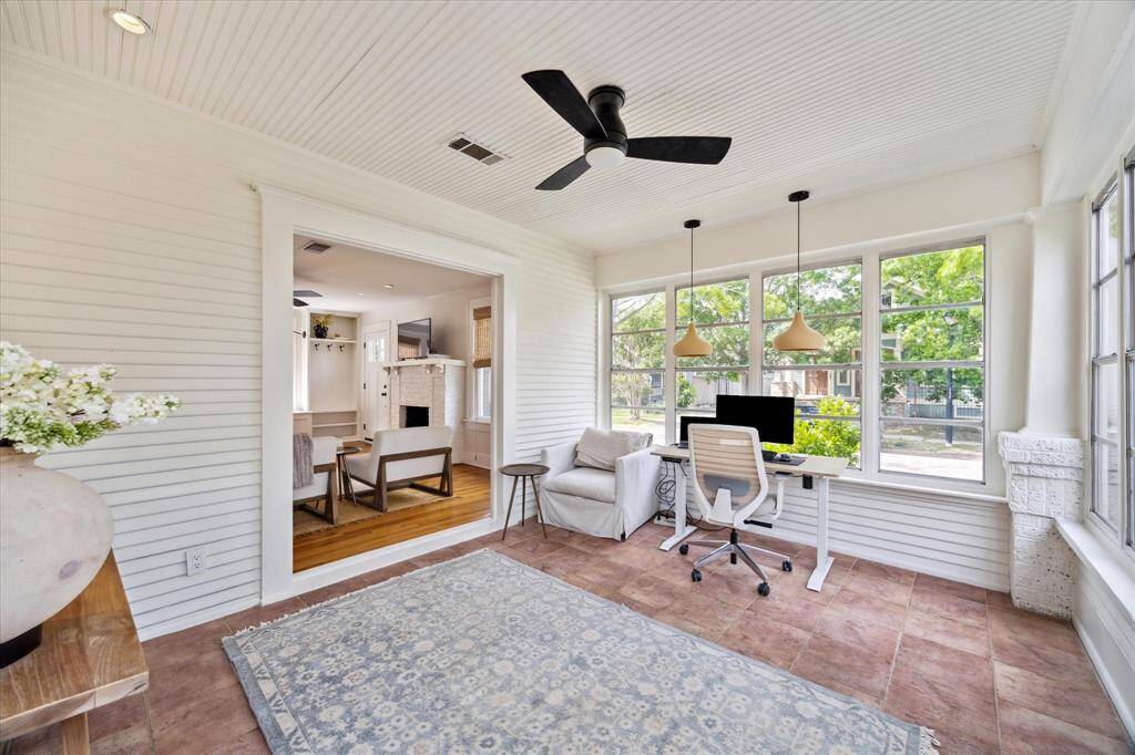 Bright sunroom utilized as a home office with large windows, offering ample natural light. Features include a ceiling fan, stylish pendant lights, and a view into the cozy living room with a fireplace. Perfect for a productive workspace.