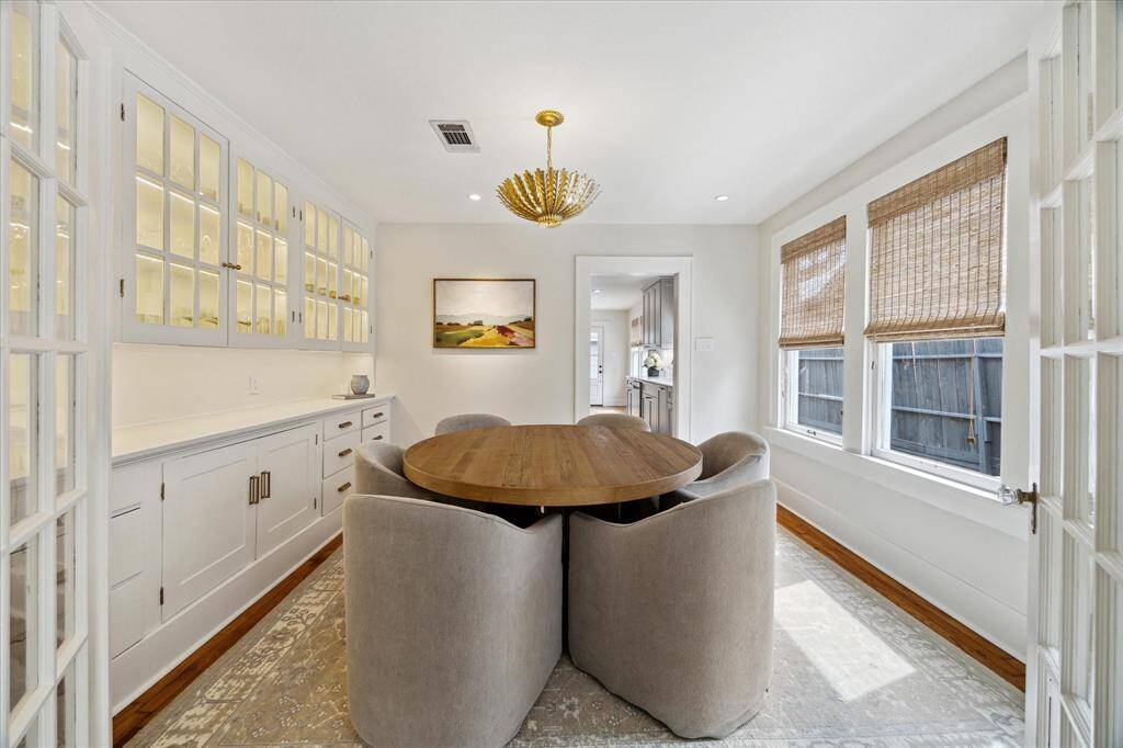 Bright dining area featuring a round wooden table with plush chairs, elegant cabinetry with glass-front doors, and large windows with bamboo shades.
