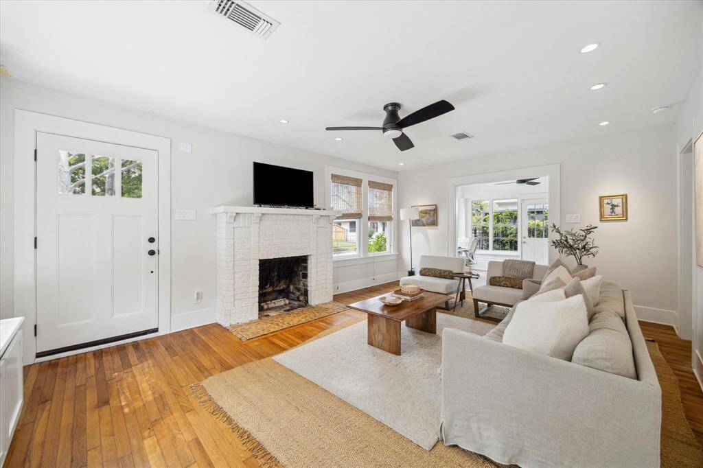 A bright and inviting living room featuring hardwood floors, a white brick fireplace, and modern ceiling fan. Large windows offer plenty of natural light.