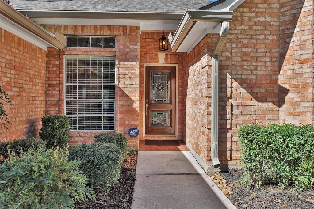 This is a welcoming entryway to a brick home featuring a large window with blinds, an ornate glass-paneled front door, and mature shrubbery. The home is equipped with a security system, as indicated by the sign.