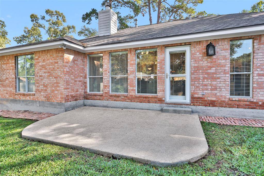 This is a single-story brick home featuring a small concrete patio area, multiple windows with blinds, and a glass-paneled back door, set against a backdrop of mature trees.