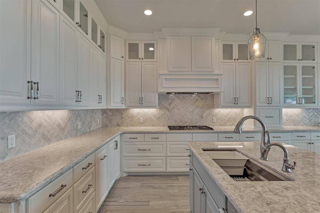 A high-end Thermador gas cooktop is paired with a custom vent hood, combining performance with designer appeal. The 42" upper cabinets, accented with glass display cabinets, add visual height and elegance. A striking herringbone-patterned backsplash brings just the right amount of texture and dimension, elevating the kitchen’s overall aesthetic.