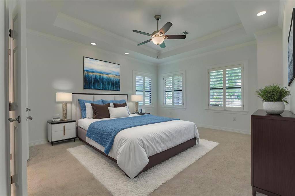The second primary bedroom is generously sized and features windows overlooking the front yard, dressed with custom shutters for added privacy and charm. Dimmable lighting on either side of the room offers versatile ambiance, while a tray ceiling enhances the sense of space and openness. Photo has been virtually staged to show furniture placement.