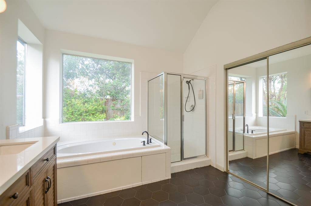 This is a spacious, well-lit bathroom featuring a large tub, separate glass-enclosed shower, dual sinks with ample counter space. The room has a clean, modern aesthetic with dark floor tiles contrasting with lighter walls.