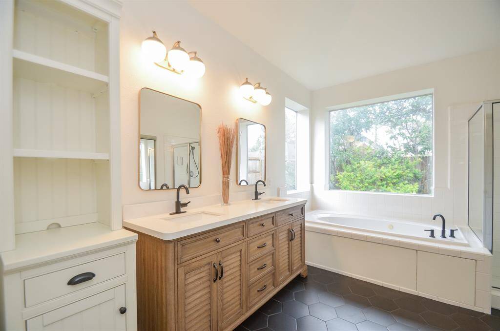 This is a bright, spacious bathroom featuring dual vanities with a mix of white and wood cabinetry, a large mirror, a built-in bathtub under a window, and a separate shower stall. The room has a clean, neutral color palette and hexagonal floor tiles.
