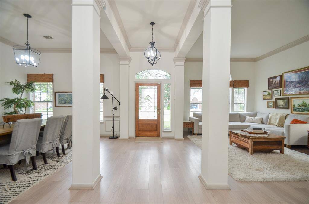 This open-concept living space features elegant white columns, high ceilings, and natural light from large windows with wooden blinds. The area is finished with light luxury vinyl plank floors and includes a cozy living area and a formal dining space. The inviting ambiance is ideal for entertaining and everyday living.