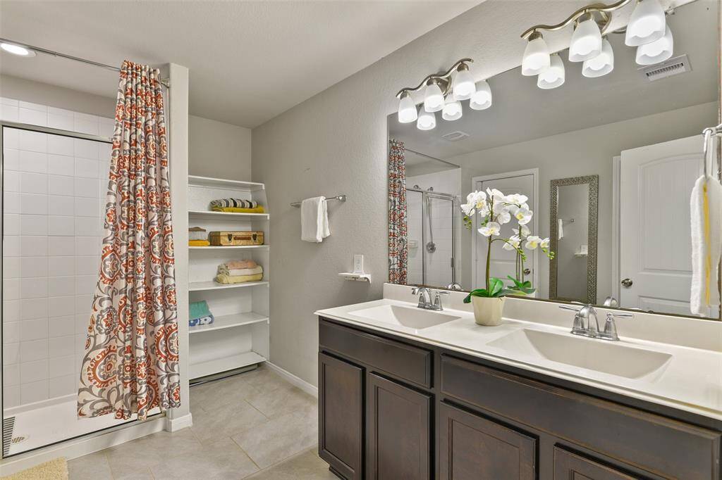 Relaxation awaits in this master bathroom with a lengthy cultured marble countertop with dual rectangular basin sinks, brushed nickel faucets, espresso-stained cabinetry, tile floors, built-in shelving, and a walk-in shower with a glass door and a tile surround.