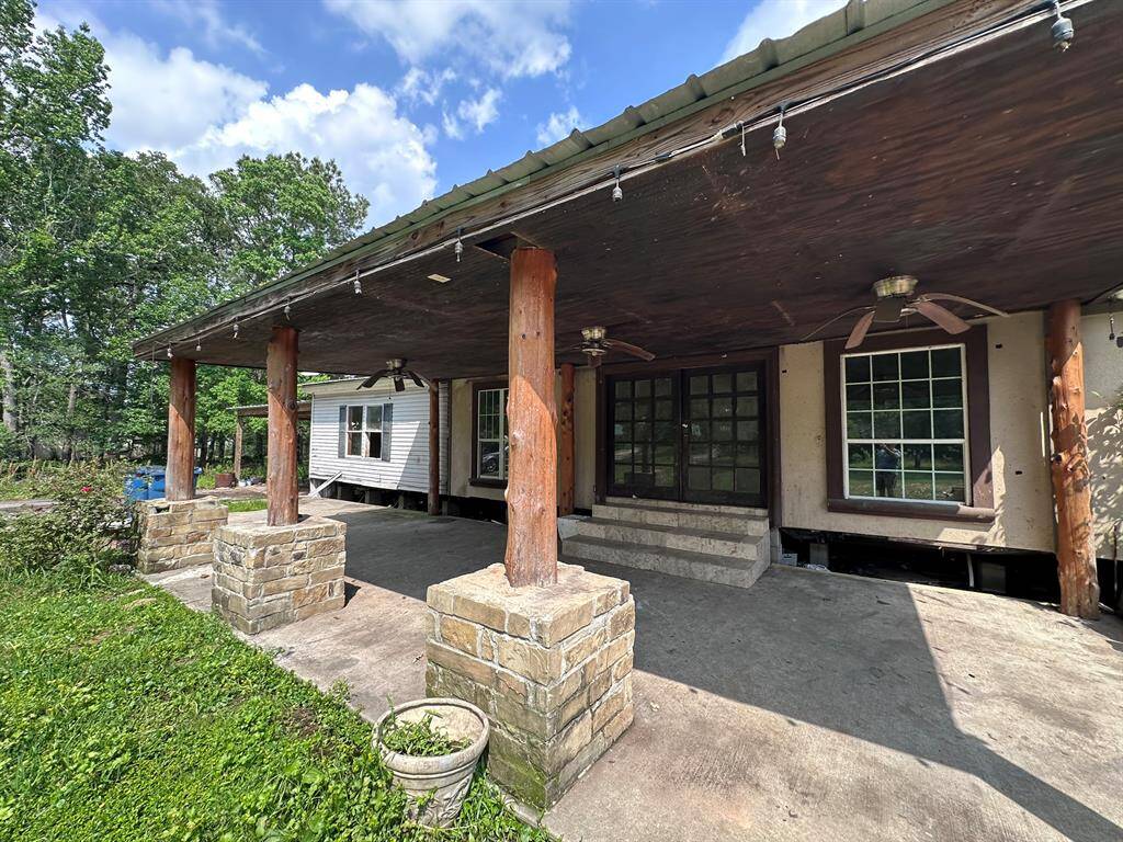 This photo showcases a rustic outdoor patio with a covered roof supported by wooden pillars set on stone bases. The area includes ceiling fans and string lights, offering a cozy space for relaxation. Lush greenery surrounds the patio, adding to the serene atmosphere.