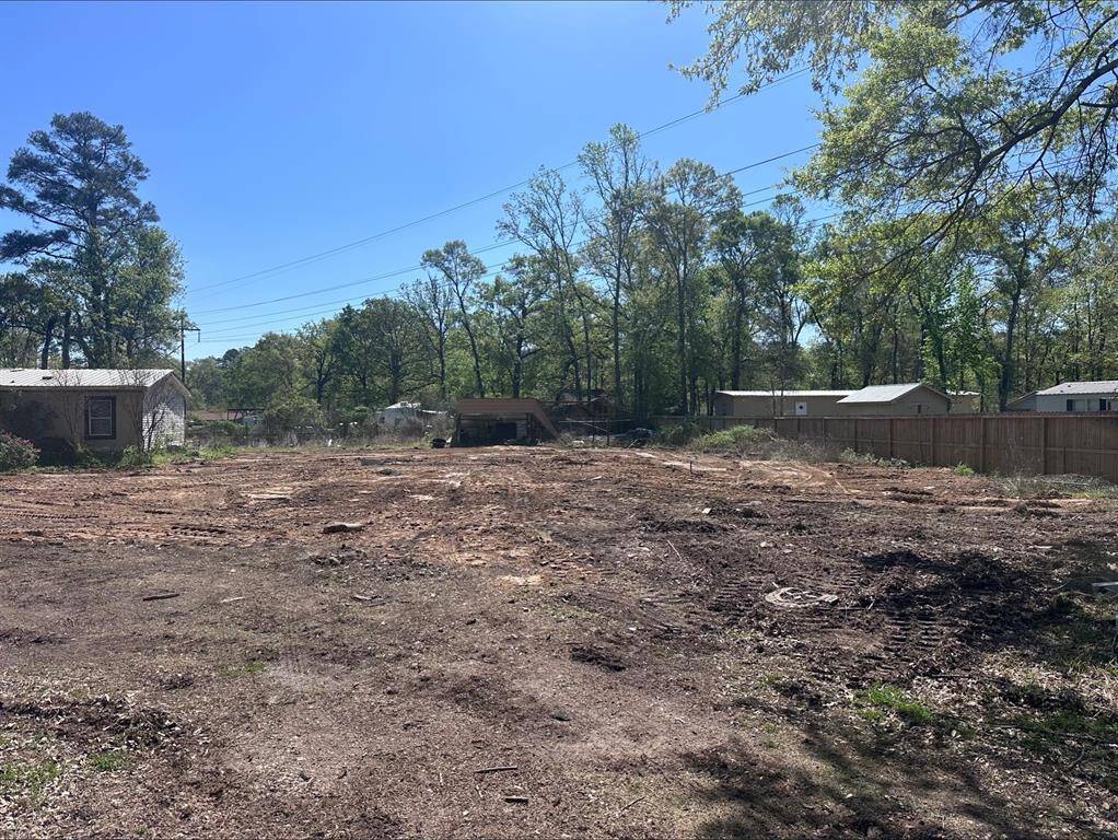 This photo shows a cleared plot of land with a few trees around the perimeter, bordered by a wooden fence. The area is flat and open, ideal for new construction, and is located in a quiet, residential setting.