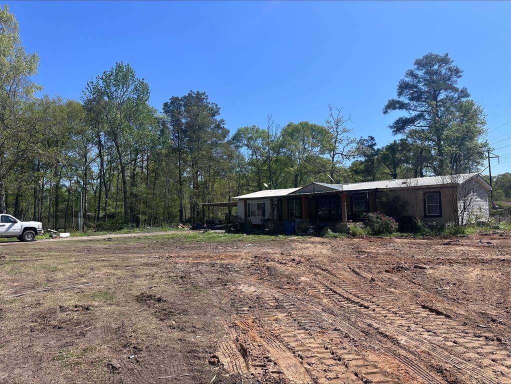This photo shows a single-story home with a metal roof situated on a spacious, partially cleared lot surrounded by tall trees. There's a truck parked nearby, and the area appears to have been recently cleared or graded, suggesting potential for landscaping or expansion.
