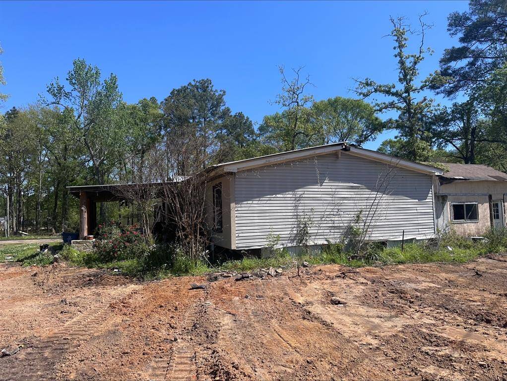 This photo shows an older mobile home with visible wear, surrounded by trees. The yard is largely bare dirt with some overgrown vegetation. It appears to need renovation and landscaping.
