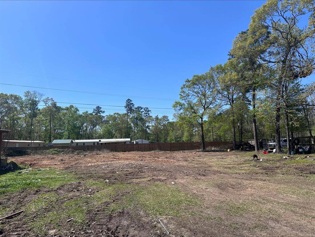 This photo shows a spacious, cleared plot of land with a few trees around the perimeter. It is bordered by a wooden fence and has a backdrop of neighboring homes and a wooded area, offering a blend of open space and privacy.