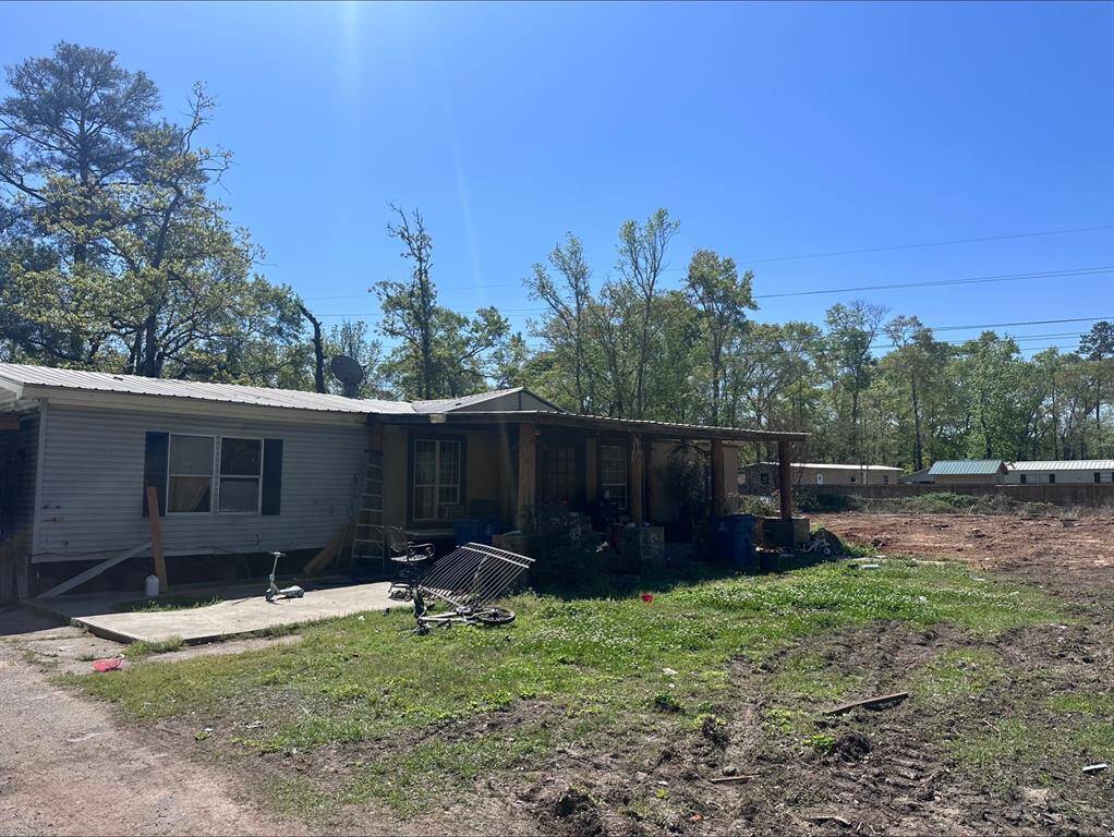 This photo shows a single-story home with a metal roof and a small covered porch. The property has a spacious, open yard with some trees in the background. The exterior appears to need some upkeep, and there is visible outdoor clutter.