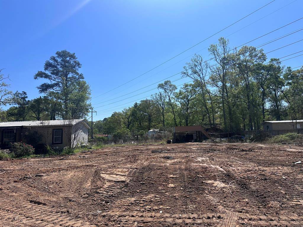 The photo shows a cleared, dirt-covered lot surrounded by trees and utility lines. Nearby are a few small structures, including a house and a shed. The area appears ready for development or landscaping.