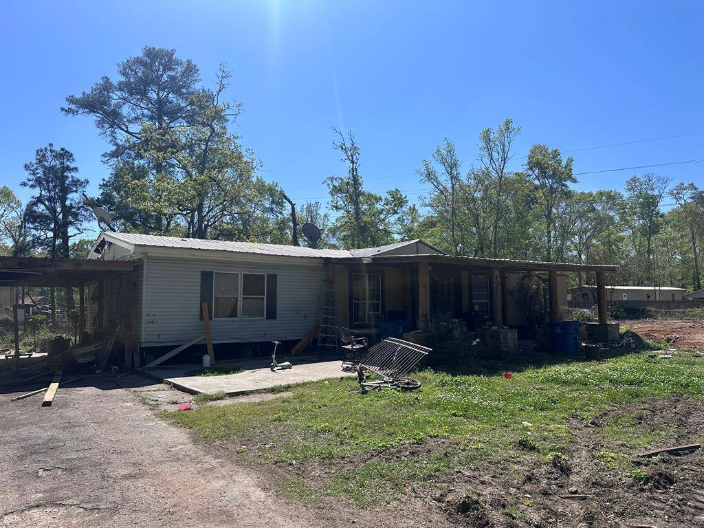 This photo shows a single-story home with a metal roof and a covered front porch. The property has a spacious yard with some scattered items and is surrounded by tall trees, providing a natural setting. The exterior may need some maintenance and updates.