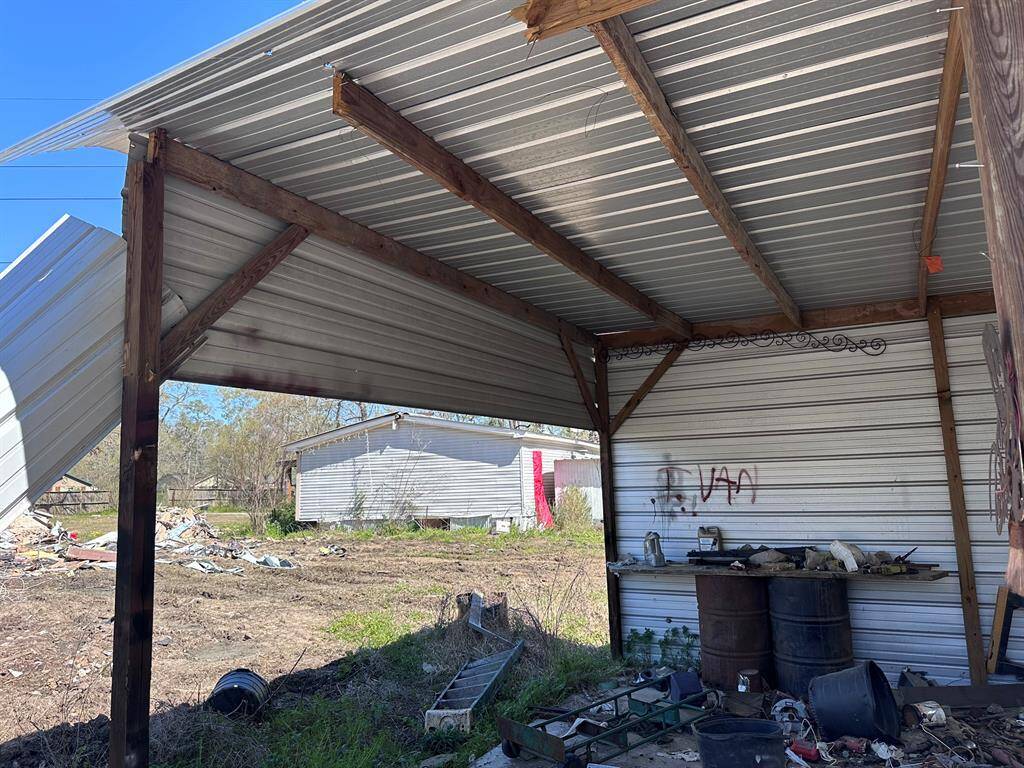 This photo shows a partially open, metal-roofed structure with wooden supports. The area is cluttered with debris and has a graffiti-marked wall. The yard appears overgrown, and a mobile home is visible in the background.