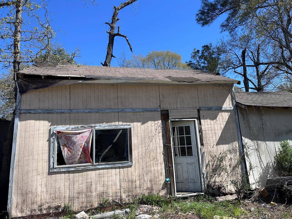 This photo shows a small, weathered structure with visible damage. The siding is worn, and the roof appears to be in disrepair. The window is broken, and the door is slightly ajar. The surrounding area is overgrown with vegetation, indicating the need for significant renovation and landscaping.
